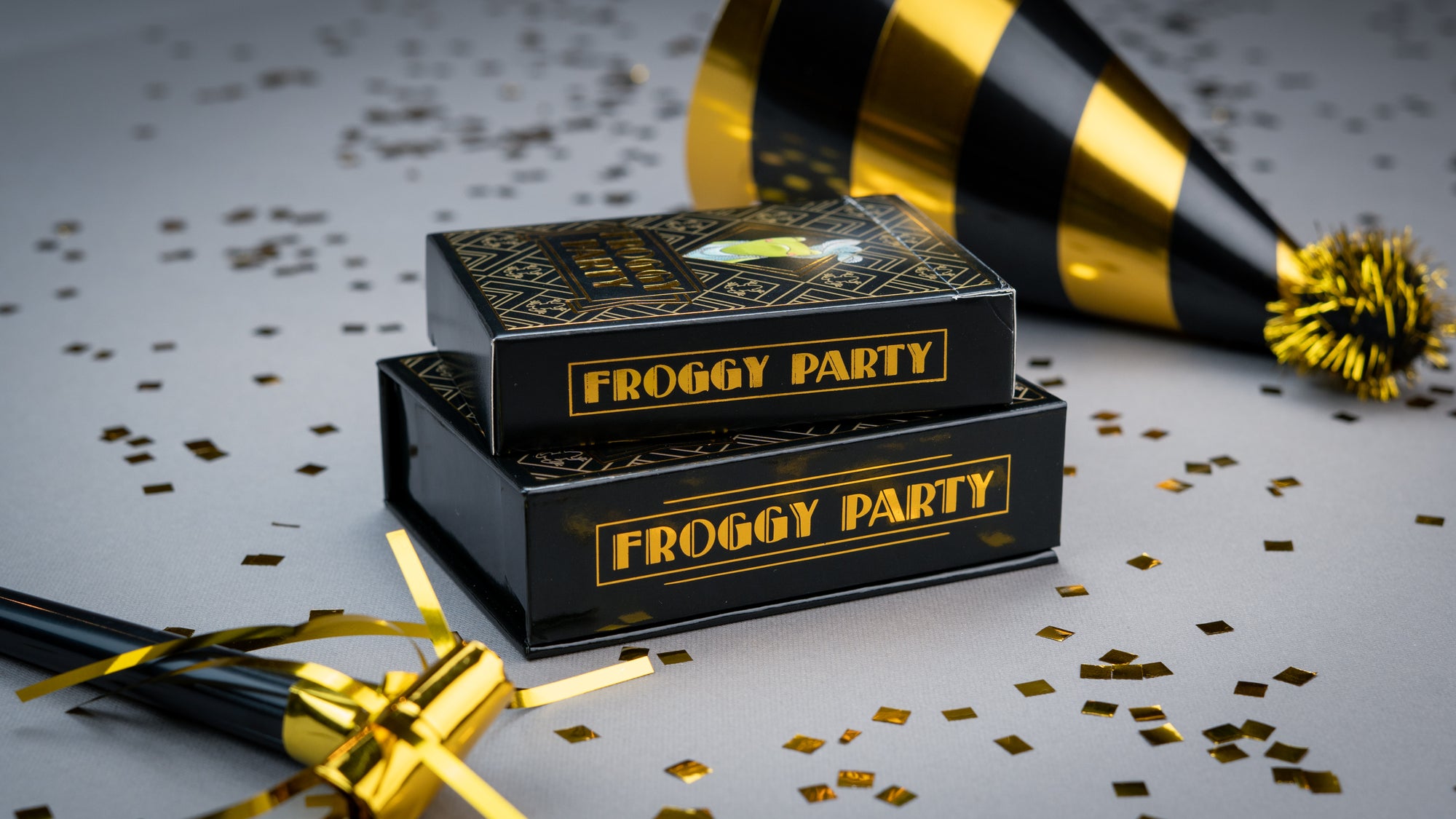 Black 'Froggy Party' boxes with gold text on a white surface with confetti and a party horn.