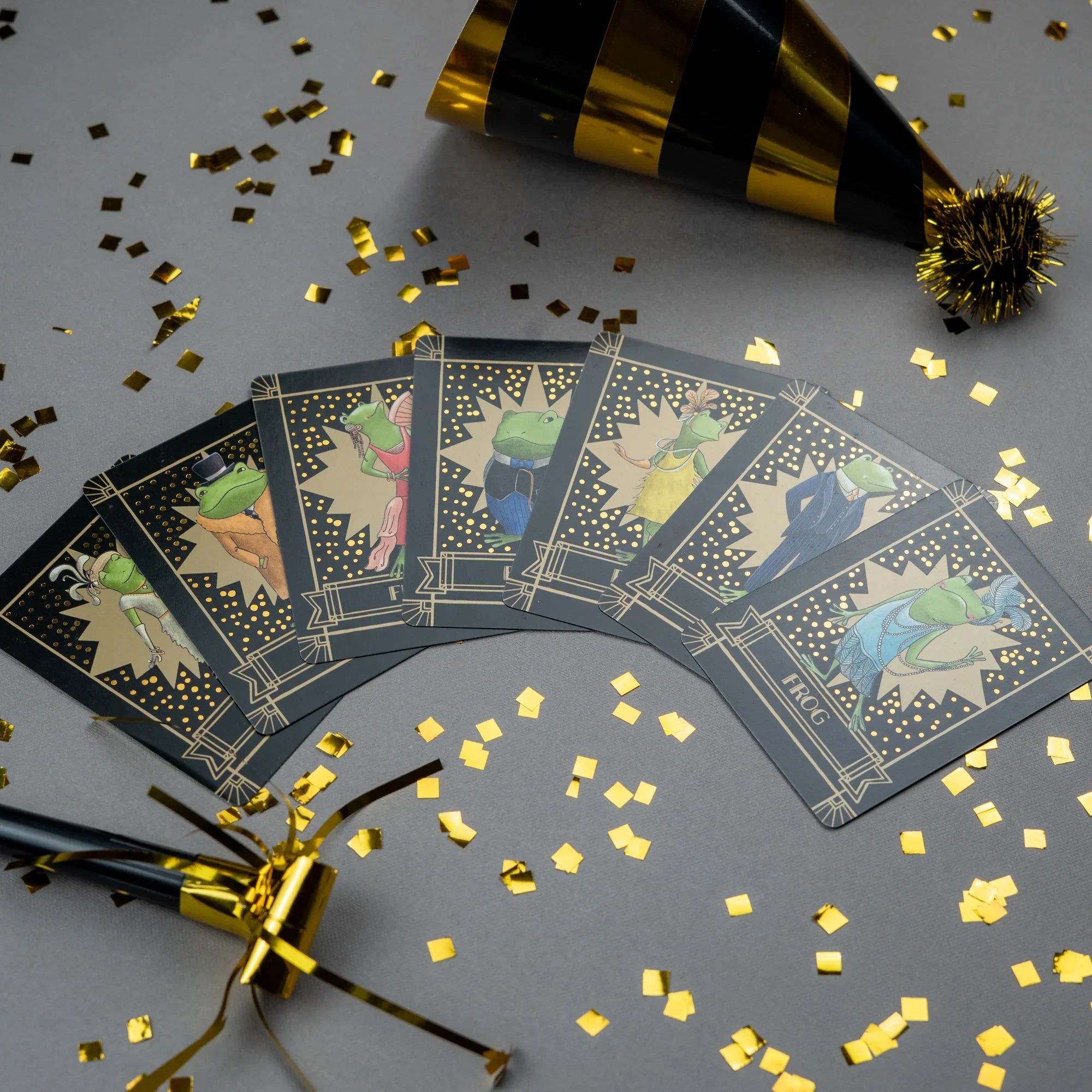 Fanned-out sparkly Frog cards with gold confetti and party hats on a gray background