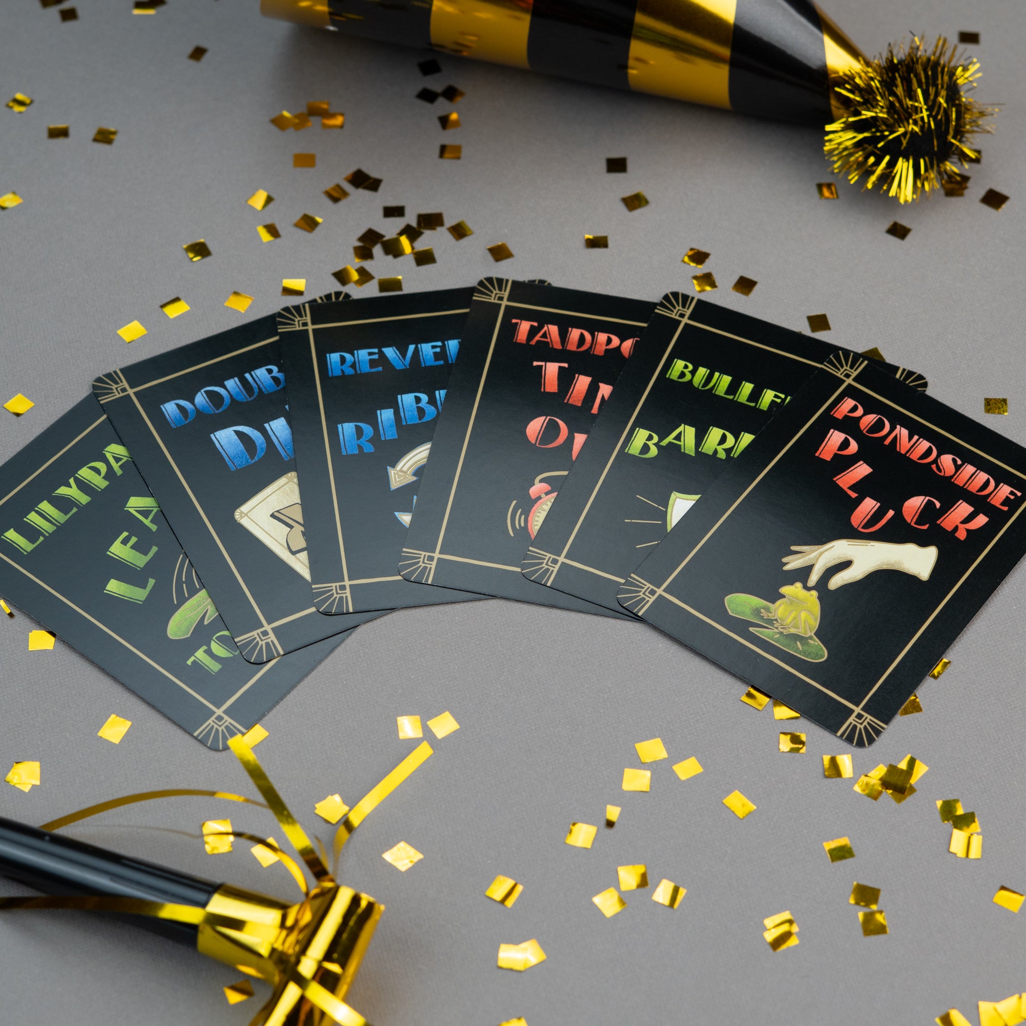 Set of gameplay cards with colorful text and designs on a gray surface with gold confetti and streamers.